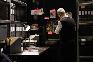 Senior police inspector looking at evidence board, analyzing victim report while working late at night at criminal case in arhive room. Elderly private detective checking criminology files photo