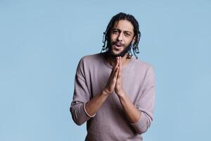 Arab man pleading with folded hands and hopeful facial expression portrait. Young adult person begging and looking at camera with pleading emotions while posing for studio shot photo