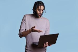 Confused arab man explaining business plan to customer in online meeting on laptop. Irritated young person holding portable computer and having conversation in video call photo