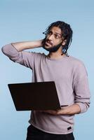 Arab programmer holding laptop, uncertain about software app development. Young person rubbing head and looking away while thinking about business report on portable computer photo