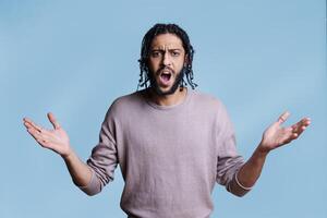 Arab man with open arms and angry facial expression looking at camera. Young adult handsome confused model with dissatisfied emotion posing for studio shot on blue background photo