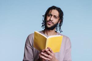 confuso árabe hombre leyendo libro de bolsillo libro y mirando a cámara con perplejo facial expresión. joven cuestionado persona participación abierto tapa blanda libro de texto y aprendizaje retrato foto