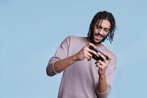 Smiling young arab man playing video game on smartphone and having fun. Cheerful person using entertaining mobile phone software application, enjoying online leisure activity photo