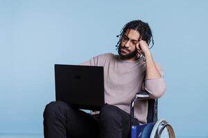 cansado árabe hombre en silla de ruedas mecanografía en ordenador portátil mientras trabajando remotamente joven agotado persona de libre dedicación software desarrollador con físico impartir analizando código en portátil computadora foto