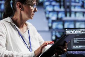 Aged system administrator writing script code in data center on laptop terminal. Skillful employee doing maintenance work in server room, updating racks software to prevent malfunctions photo