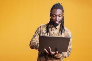 African american man holding laptop computer browsing webpages on internet, standing in studio over yellow background. Young adult using portable device to navigate on social media photo