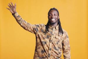 African american man waving with palm at camera, smiling person being friendly in studio. Cheerful person doing salute gesture to greet people while standing over yellow background photo