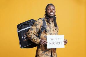 Takeout delivery employee with thermal backpack holding hiring application sign for courier position. African american person wanting perfect candidate for career opportunity in studio photo