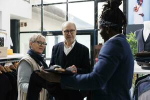 Elderly couple discussing shirt fabric with showroom worker, buying trendy clothes for formal wear in modern boutique. Senior man looking at new fashion collection in shopping centre. Fashion concept photo