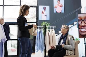 Store employee helping senior customer with new collection, trying fashionable shoes in shopping center. Elderly client buying stylish clothes and merchandise, department store small business. photo