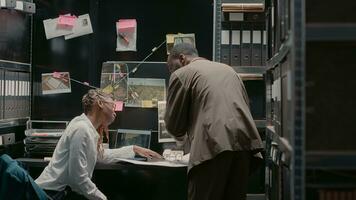 African american detectives looking at photos in police archive, doing research to solve criminal case. Inspectors examining physical evidence to uncover details on investigation crime board.