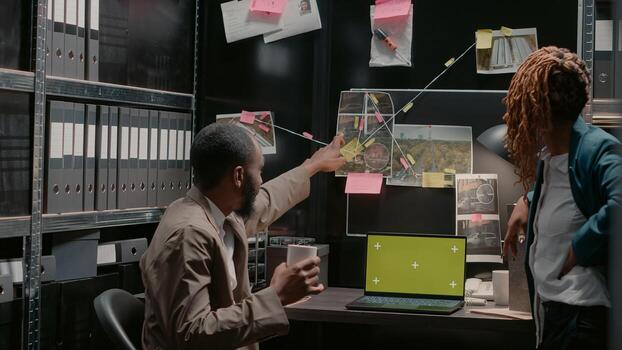 Two inspectors using greenscreen on laptop at desk, analyzing case reports in police archive files. Private detectives team looking at isolated display with blank copyspace on screen. photo