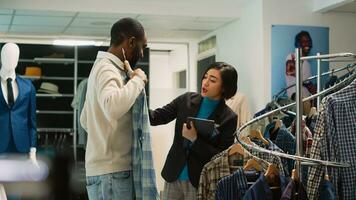 Diverse people examining clothes on racks to do shopping, woman working as store assistant in shopping center to help man choose shirts. Client looking at fashion merchandise. Handheld shot. photo