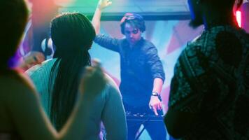 Group of friends dancing in the club, enjoying live performance show with DJ mixing station on nightclub stage. Young people partying together on dance floor with disco lights. Tripod shot. photo