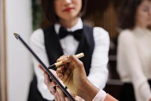 Guest filling in papers at front desk, talking to hotel employee about accommodation in lobby. Young woman doing check in process at reception, writing registration papers. Close up. photo