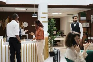 Couple checking in at hotel reception, asking receptionist about accommodation and room reservation. People in lobby preparing to fill in registration forms, receiving luxury service. photo