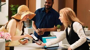 Diverse couple using mobile nfc payment with smartphone next to pos terminal, paying for room reservation at tropical summer resort. Tourists doign check in at front desk reception. photo