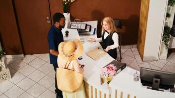 diverso Pareja haciendo cheque en con recepcionista, hotel conserje saludo turistas de viaje en verano vacaciones. invitados llegando a frente escritorio en vestíbulo, habitación reserva. Mano disparo. foto