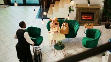 Bellhop offering services to woman in lounge area, waiting to do room check in at seaside hotel. Tourist sitting on couch and talking to hotel concierge, summer clothes. Handheld shot. photo