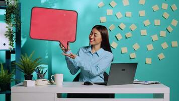 Happy cheerful asian businesswoman holding red speech bubble sign of empty space for text and showing ok hand gesture. Smiling employee presenting thought bubble cardboard in colourful workplace photo