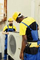 Precise serviceman and african american colleague installing new external hvac system for client. Meticulous wiremen optimizing new air conditioner performance, ensuring it operates at productivity photo