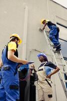 Competent wiremen diverse coworkers commissioned for external air conditioner routine check up, writing findings on clipboard. Professionals doing hvac system investigation of damaged parts photo