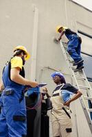 Competent wiremen diverse coworkers commissioned for external air conditioner routine check up, writing findings on clipboard. Professionals doing hvac system investigation of damaged parts photo