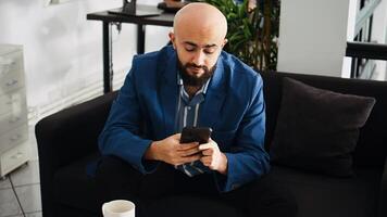 Startup employee works on presentation, using smartphone to analyze statistics in coworking space. Entrepreneur reviewing data at workstation, planning project in open floor plan. photo