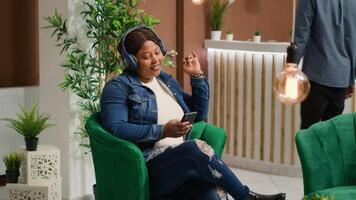 Traveller listening to music in reception lobby, having fun with audio headset and songs on smartphone. Woman waiting to do check in process at luxury hotel, sitting in lounge area on holiday. photo