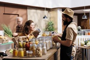 Woman shopping in zero waste grocery store, looking at bulk products in reusable packaging while being assisted by helpful clerk. Customer in local neighborhood picking ethically produced food photo