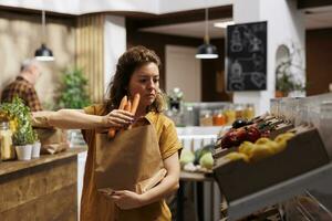 Environmentally responsible shopper buying ecofriendly groceries in zero waste store using paper bags to minimize plastic usage. Green living customer looking for healthy chemicals free vegetables photo