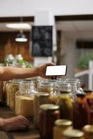 Client in zero waste shop using mockup mobile phone to analyze pantry staples. Vegan customer thoroughly checking local supermarket food items are allergens free with isolated screen smartphone photo