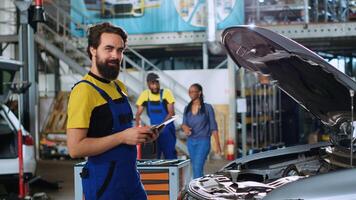 Portrait of smiling skillful mechanic in auto repair shop doing car annual checkup using tablet, looking for damages. Cheerful repairman at work checking to see if vehicle components need replacement photo