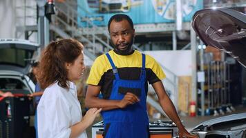 Certified worker in repair shop showing client damages found inside car after inspection. African american professional telling woman vehicle diagnostic after checking for defective parts photo