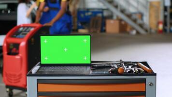 Green screen laptop placed on working bench in busy garage next to professional tools while mechanics walk around in blurry background. Chroma key device in auto repair shop, close up photo