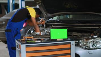 Engineer in car service uses torque wrench to secure bolts inside vehicle with green screen tablet in front. Expert utilizes professional tools to mend out of order automobile next to mockup device photo