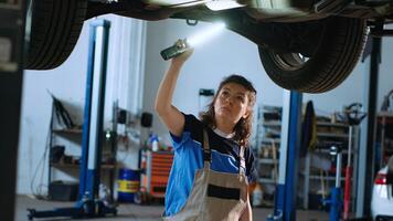 Mechanic underneath vehicle on overhead lift in garage, checking parts during annual checkup. Car service professional using work light to make sure automotive underbody is in proper condition photo