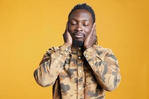 Man with close eyes covering ears with palms, doing three wise monkeys gesture in front of camera over yellow background. African american person not listening to noise and not speaking in studio photo