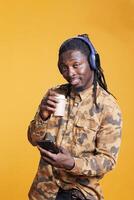 Positive smiling adult holding cup of coffee, holding smarphone scrolling to social media during lesiure time in studio. African american man searching information on internet using mobile phone photo