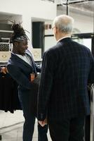 Store employee helping client choosing formal suit in showroom, african american worker helping customer to buy modern clothes. Senior man buying fashionable merchandise in modern boutique photo