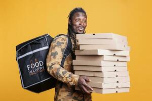 pizzería entrega empleado participación cartulina apilar lleno con pizza, entregando para llevar comida orden a cliente durante almuerzo tiempo. africano americano repartidor que lleva térmico mochila en estudio foto