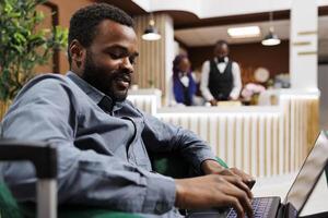 Smiling young African American man entrepreneur using laptop while waiting with suitcase in hotel lobby. Businessman working remotely during business trip, looking at computer screen photo