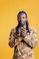 Successful positive man standing with palms together, asking for something in studio over yellow background. African american person celebrating succes, applauding people after job achievement photo