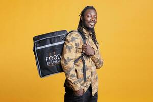 African american deliveryman carrying food delivery thermal backpack, standing in studio over yellow background. Restaurant courier deliverying healthy meal to customers during lunch time photo