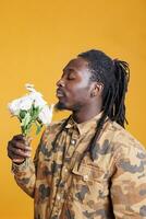 Portrait of joyful man enjoying white roses smell, posing with flowes for valentine s day in studio over yellow background. African american romantic young adult celebrating love holiday photo