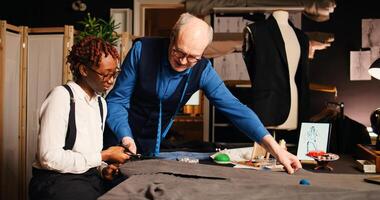 Trainee and craftsman cutting fabric, teaching the process to design custom made tailored clothes. Female beginner using scissors and preparing garment to design clothing with old tailor. photo