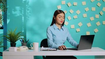 Happy cheerful asian businesswoman imputing data at modern bright creative office desk over blue studio background. Smiling employee imputing data in relaxed administration workplace photo