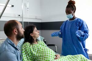 africano americano cirujano preparando embarazada mujer para cirugía, explicando parto proceso en hospital pabellón. paciente con el embarazo acostado en cama teniendo contracciones consiguiendo dentro labor foto