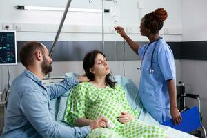 africano americano enfermero comprobación embarazada mujer iv goteo, preparando para médico cirugía en hospital pabellón. paciente con el embarazo acostado en cama, que se discute con marido acerca de paternidad foto