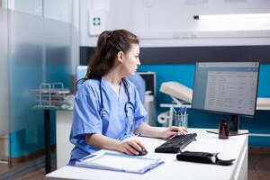 caucásico enfermero trabajando a escritorio, insertando datos en cita mesa lista. concentrado adulto mujer trabajador vistiendo matorrales y estetoscopio en limpio, moderno médico oficina, operando computadora foto